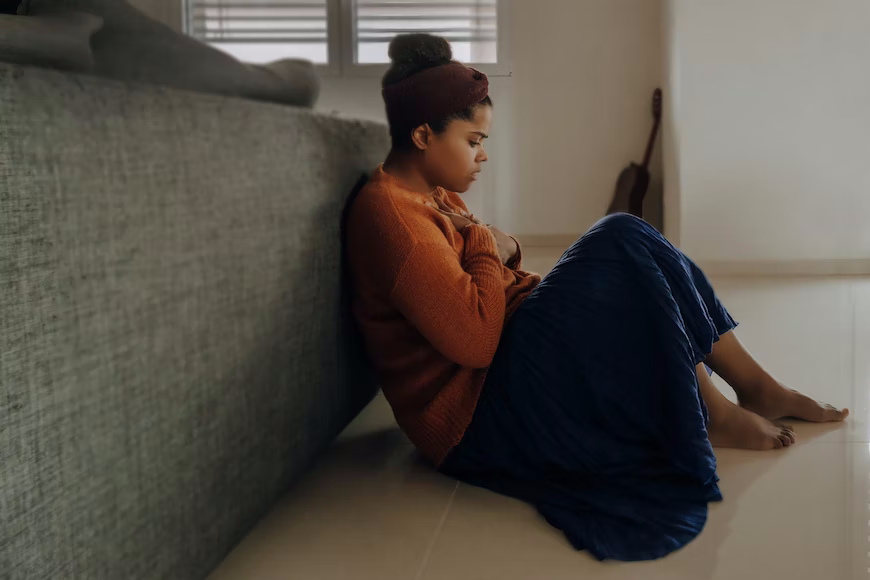 A person in an orange jumper and long blue skirt sitting on the floor holding their chest