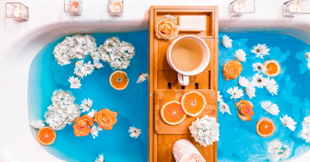 A bathtub filled with blue water, upon which are floating white and orange flowers and slices of orange. There are candles on the rim of the tub, and a caddy on top of it, on which are placed a bar of soap, a white and an orange flower, two slices of orange, a tealight holder, and a mug of tea.