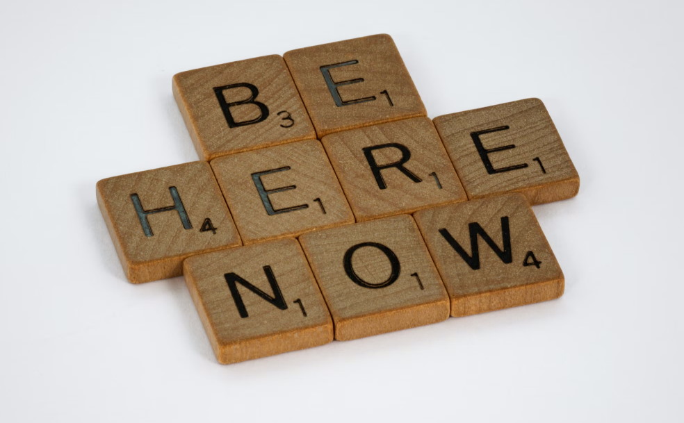 wooden scrabble letters spelling out 'be here now'