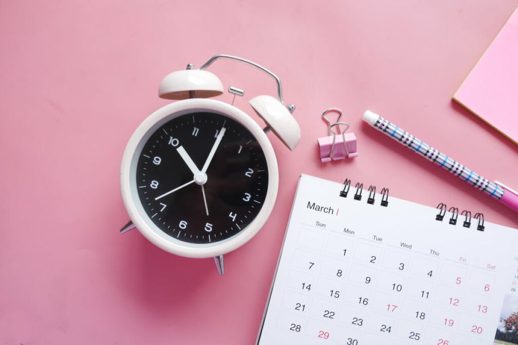 A clock, calendar and pen resting on a pink surface
