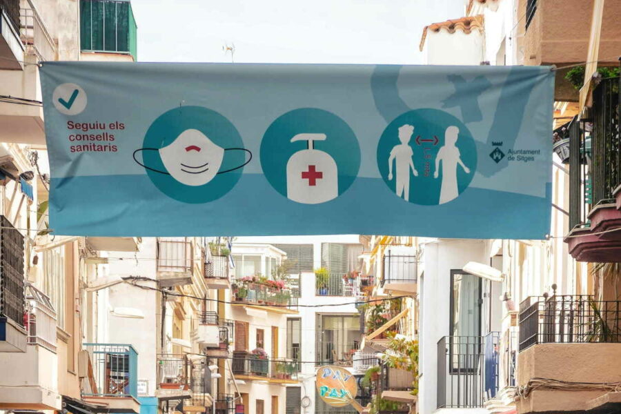 Banner above a street in Sitges, Spain, featuring icons symbolising a face mask, hand sanitiser and social distance, and text in Catalan