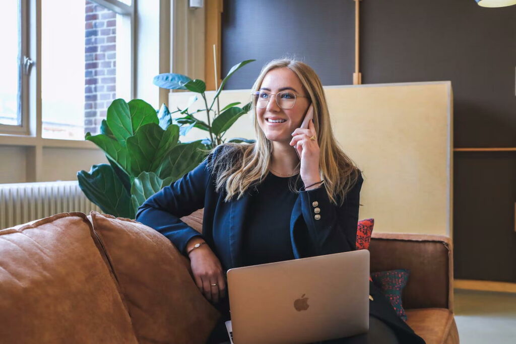 Woman with long blonde hair and glasses smiling while on phone with a Macbook on her lap. 