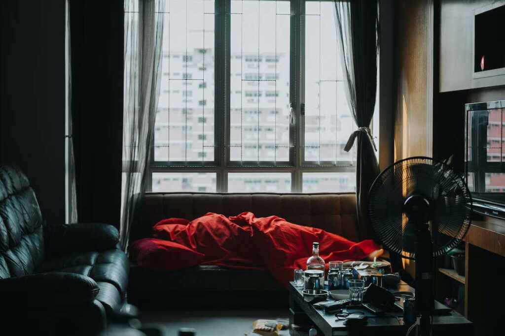 A person wrapped in a red blanket lying on the sofa in a messy room with a coffee table, fan and two sofas