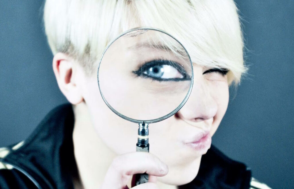 Person with short blonde hair looking through magnifying glass, making their blue eye appear larger
