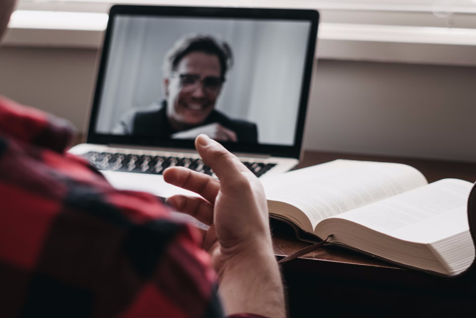 A person at a table with an open book and a laptop screen displaying the face of a person they are talking to via video chat
