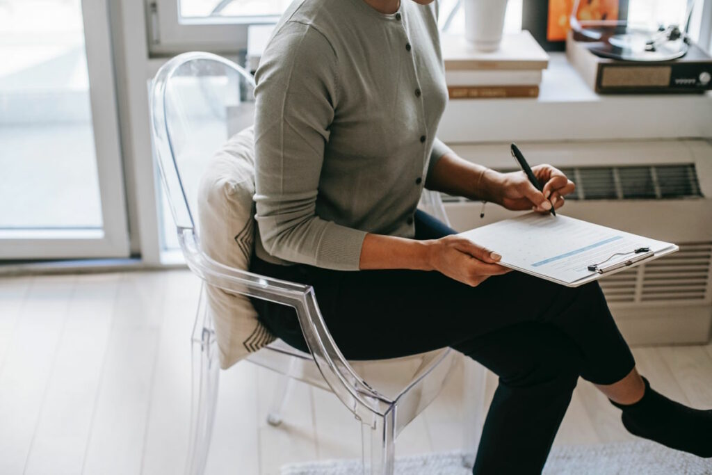 Photo of a person sat on a clear plastic chair holding a pen and clipboard, with the person's head out of shot