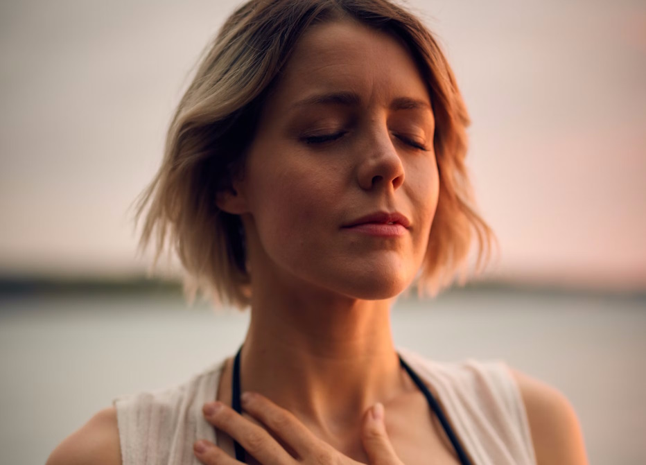 A woman in a white top focusing on her breathing, with her eyes closed and one hand on her chest.