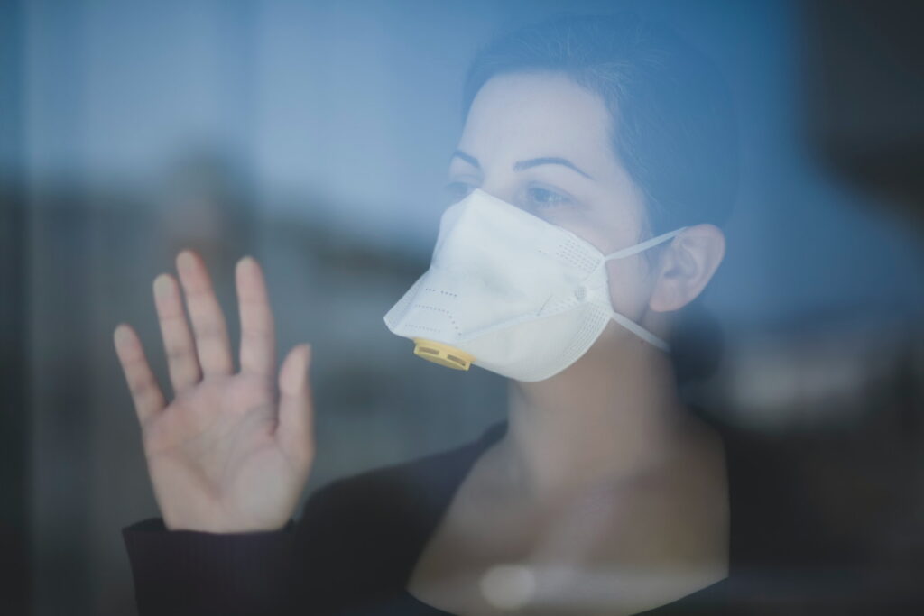 A woman wearing a face mask stood behind a window, staring into the distance and with her hand pressed against the glass