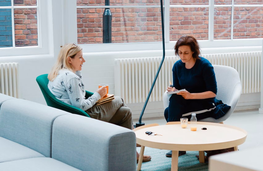 Two women in conversation, one writing on a notepad, sat in armchairs behind small round table 